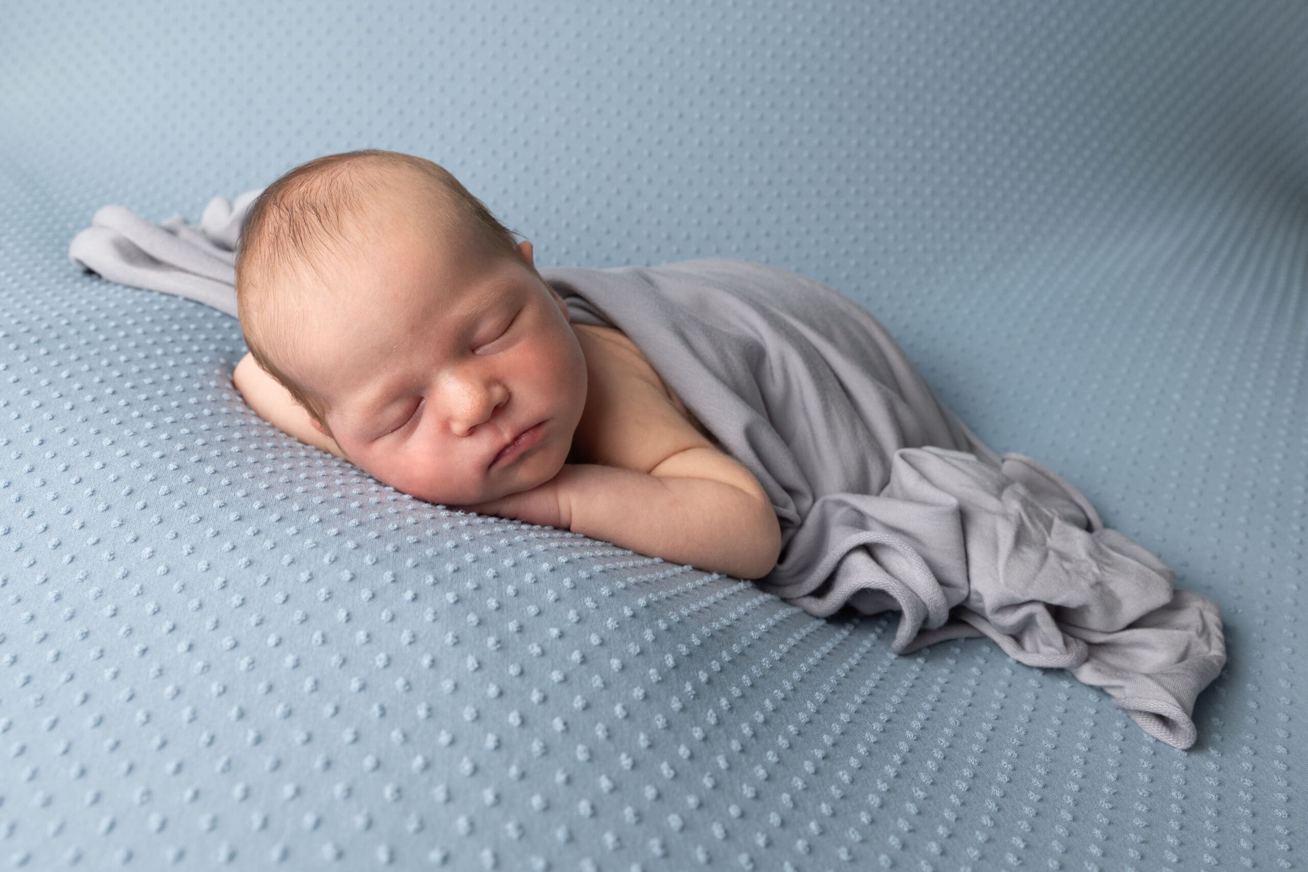 A peacefully sleeping newborn baby, wrapped in a soft grey blanket, resting on a blue textured blanket. The baby is lying on their side with their head slightly tilted, showcasing a calm and serene expression. The setting creates a cosy and comforting atmosphere, perfect for capturing a precious moment.