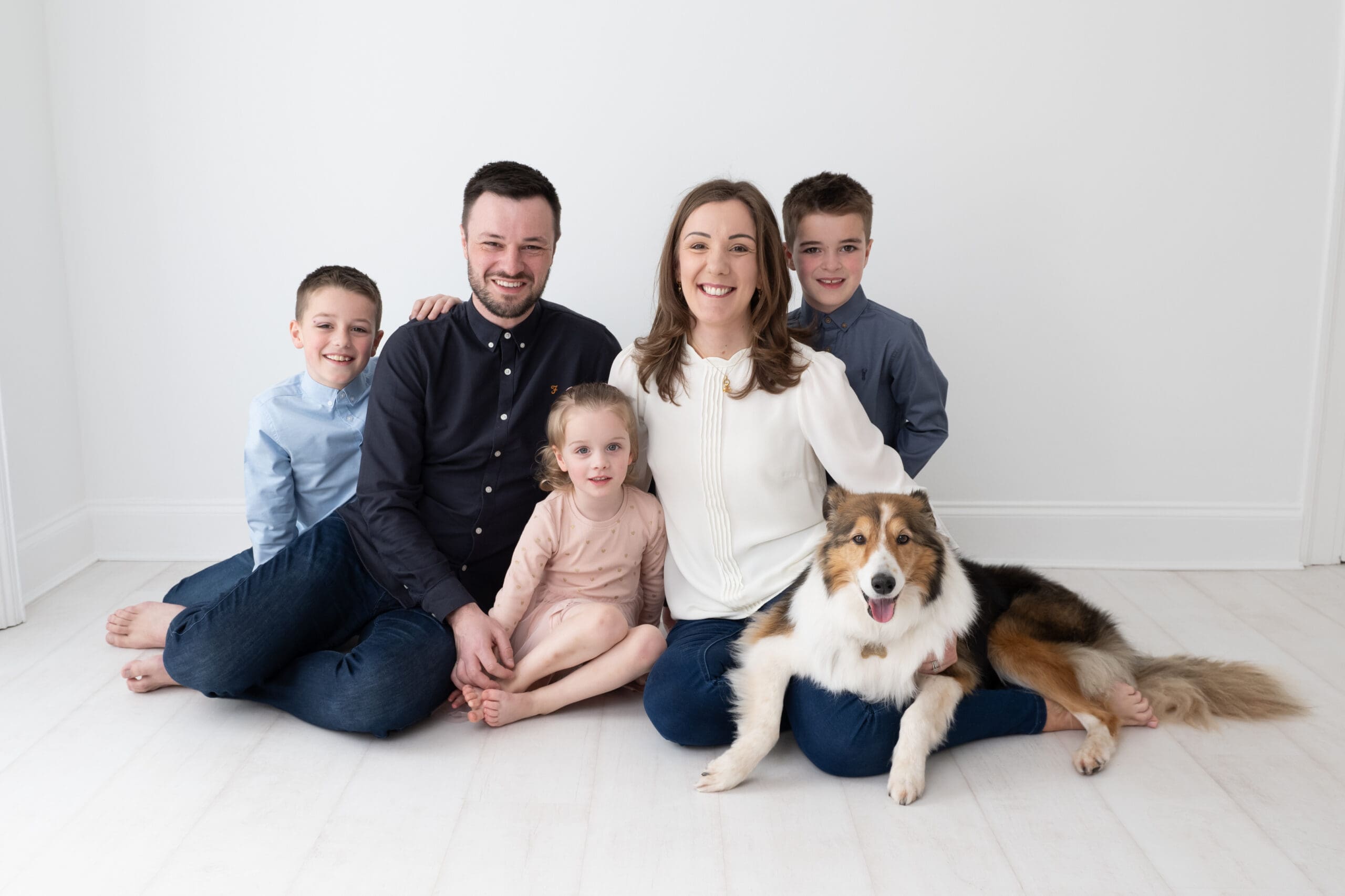 A happy family of five, including two young boys, a toddler girl, and their parents, sitting barefoot on a white floor against a bright, minimal backdrop. The family dog, a friendly Collie, sits beside them, adding a warm touch to the family portrait. Everyone is dressed in coordinated, casual outfits, with the boys in blue shirts, the father in a dark shirt, and the mother in a white blouse. The family is smiling, creating a welcoming and joyful atmosphere.