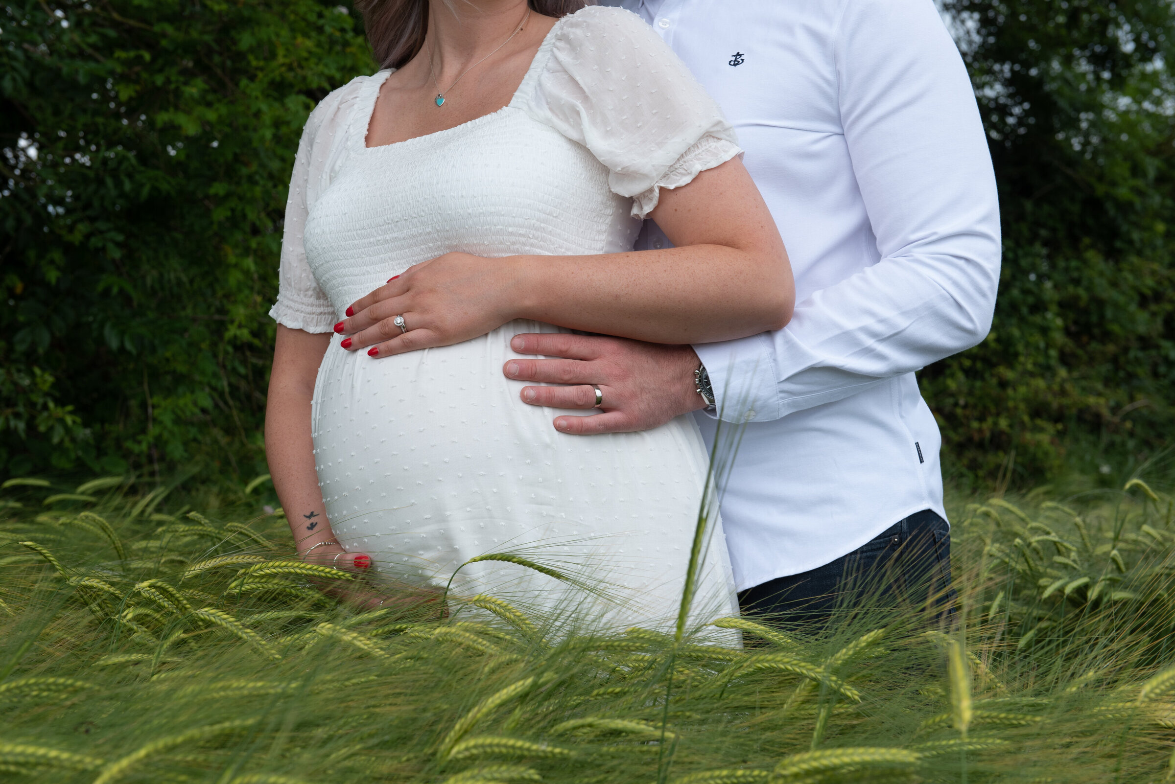 Expecting parents standing in a lush green field, gently holding the mother's pregnant belly. The mother wears a white dress with delicate puffed sleeves, while the father stands behind her, both resting their hands on the baby bump. Their wedding rings are visible, adding a personal touch to the image, which radiates love and anticipation.